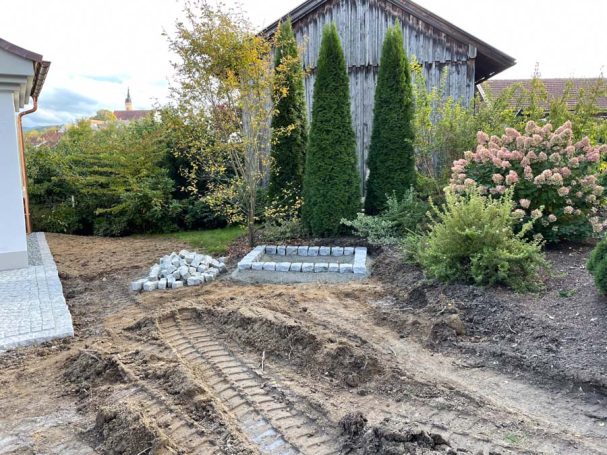 Gartenbereich mit gegrabener Erde, zwei Tannen und einem Haus im Hintergrund.