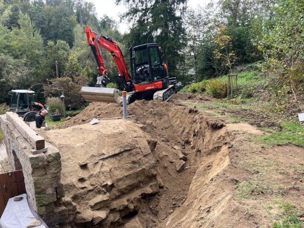 Bagger arbeitet im Garten an einem ausgehobenen Graben umgeben von Bäumen und Pflanzen.