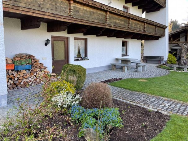 Gepflegter Garten mit Sitzgelegenheit, Holzlager und bunten Pflanzen vor einem Haus.