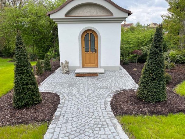Ein gepflasterter Weg führt zu einer weißen Kapelle, umgeben von Nadelbäumen.