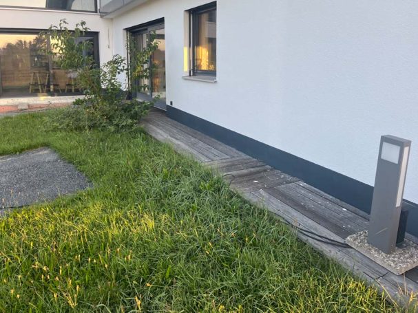 Grüner Rasen neben einer modernen Wand mit Fenstern und einem Weg, der zum Haus führt.