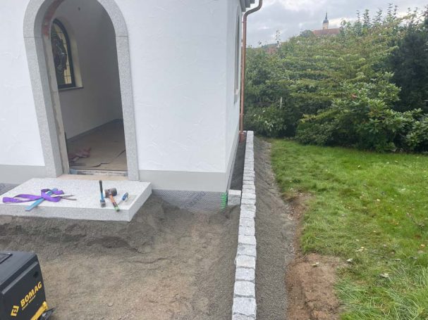 Baustelle mit unvollständigem Pflaster an einem Gebäudeeingang und angrenzendem Grünbereich.