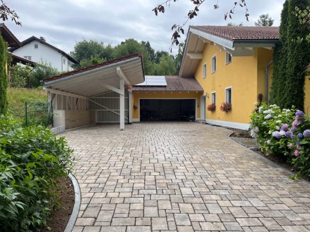 Gelbe Villa mit Carport und gepflastertem Vorplatz, umgeben von grünen Pflanzen.