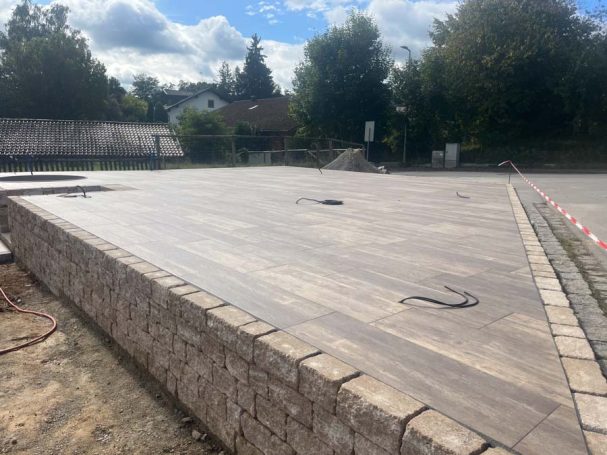 Holz-ähnlicher Bodenbelag auf einem erhöhten Areal mit Steinmauer und bewaldetem Hintergrund.