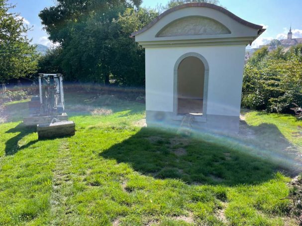 Weißes Bauwerk mit Rundbogen, umgeben von grünem Gras und Bäumen.