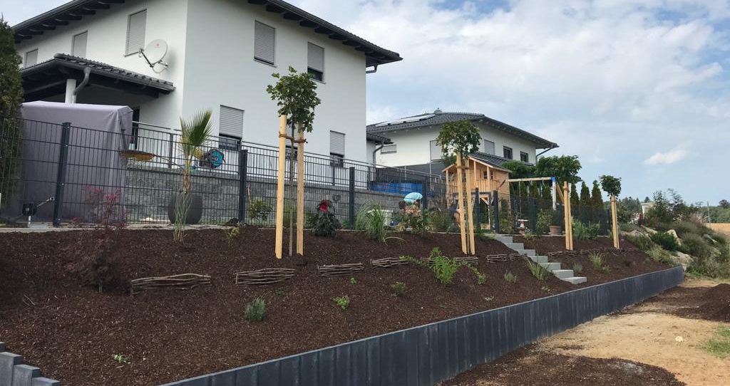 Gartenlandschaft mit Bäumen, Sträuchern und zwei modernen Wohnhäusern im Hintergrund.