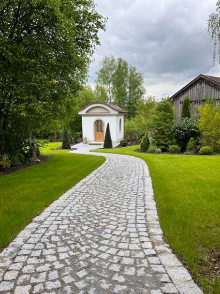 Weg aus Pflastersteinen zu einem kleinen weißen Häuschen in einer grünen Landschaft.