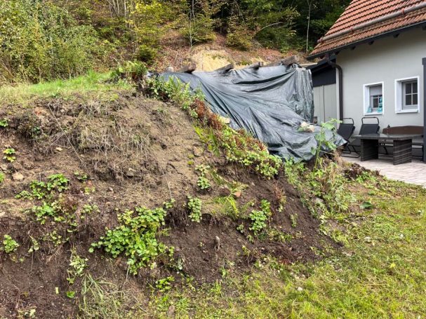 Erdhügel mit schwarzer Folie, umgeben von Gras und kleinen Pflanzen, neben einem Gebäude.