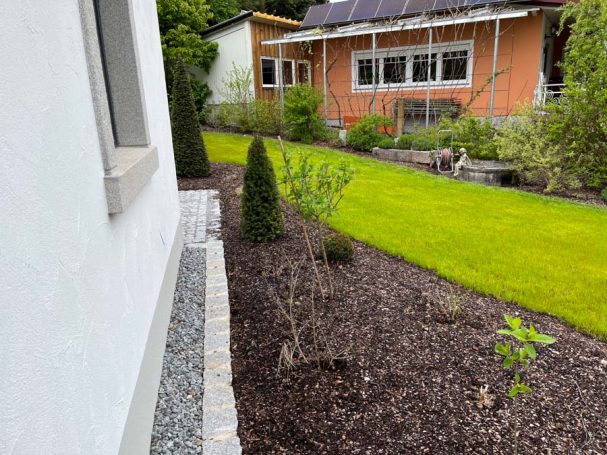 Gartenansicht mit Kies, Büschen und Rasenfläche hinter einem weißen Gebäude.