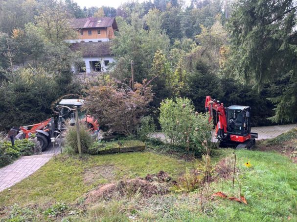 Baugeräte stehen in einem Garten mit Bäumen und einem Wohnhaus im Hintergrund.