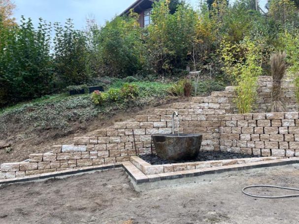 Steinmauer mit Pflanzflächen und einer runden Steinwanne in einer Gartenlandschaft.