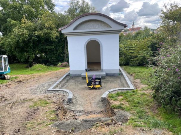 Weißes Gartenhaus mit Portal, umgeben von einem rohen Weg und grüner Vegetation.