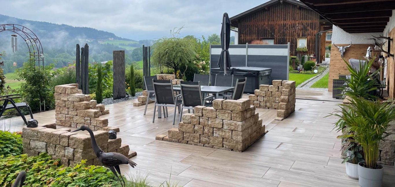 Terrasse mit Steingärten, Tisch und Stühlen, umgeben von grüner Landschaft und Bergen.