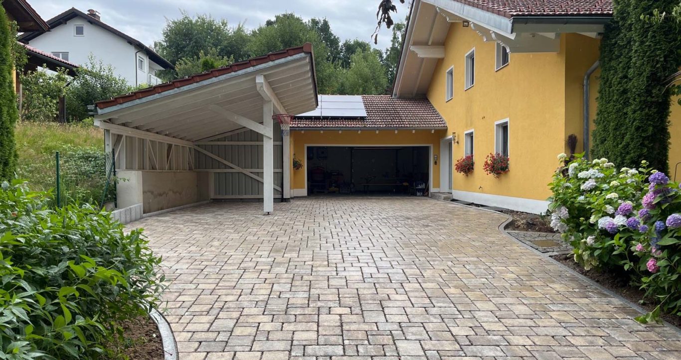 Gelbes Haus mit Carport, gepflasterter Einfahrt und Gartenpflanzen im Vordergrund.