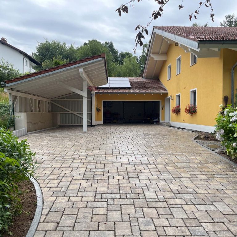 Gelbes Haus mit überdachtem Parkplatz und gepflasterter Auffahrt, umgeben von Pflanzen.