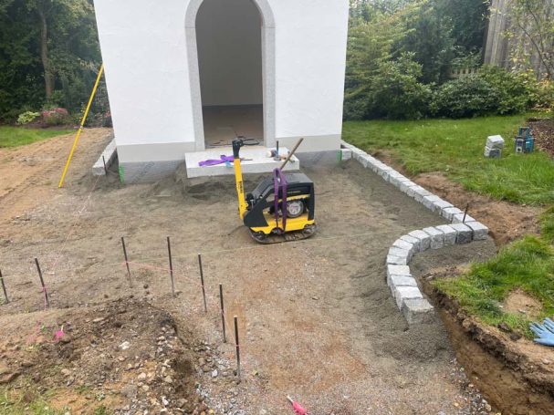 Baustelle mit Rüttelplatte und frisch gegossenem Beton um ein Gebäude.