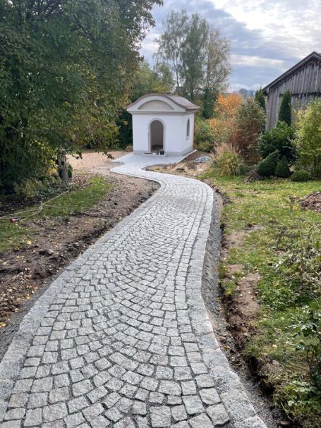 Kopfsteinpflasterweg mit kleiner Kapelle und umgebendem Grün.