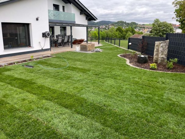 Rasenfläche mit gestreiftem Grün, umgeben von Bepflanzungen und einem modernen Haus.