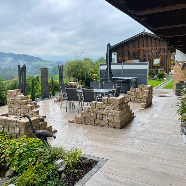 Terrasse mit Möbeln, Steinmauer und Bergblick unter bewölktem Himmel.