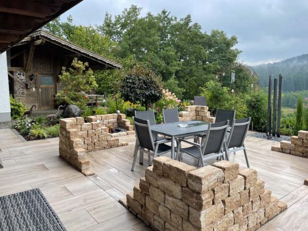 Terrasse mit Gartenmöbeln und Holzstapeln, umgeben von Bäumen und Bergen.