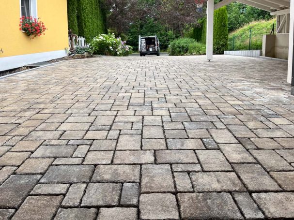 Gestampfter Pflasterboden mit hellbraunen Steinen, umgeben von Pflanzen und einer gelben Wand.