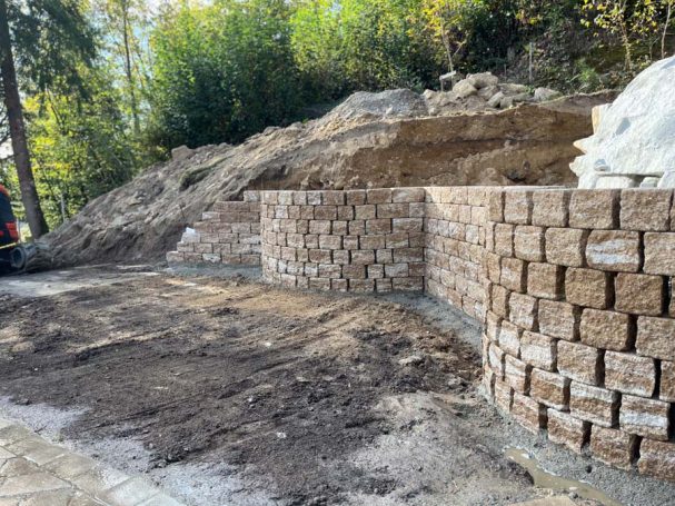 Mauer aus Flusssteinen, umgeben von unbefestigtem Boden und Bäumen im Hintergrund.