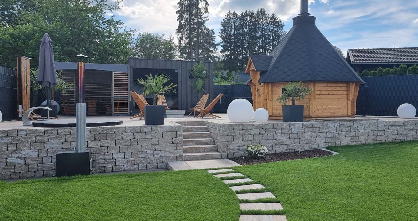 Gepflegter Garten mit Steinterrasse, Holzhaus, Pflanzen und Sitzgelegenheiten.