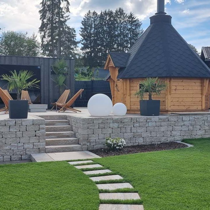 Holzhütte mit schwarzem Dach, umgeben von Rasen, Steinen und modernen Möbeln im Garten.