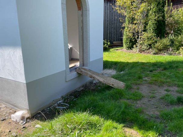 Holzbrett führt von einer Tür ins Freie, umgeben von Rasen und Büschen.