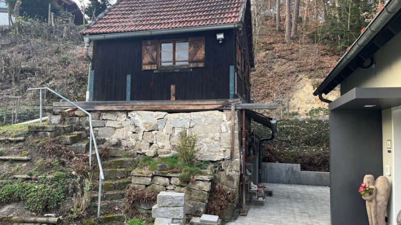 Kleines schwarzes Haus mit Fenster und Terrasse, umgeben von Steinen und Natur.
