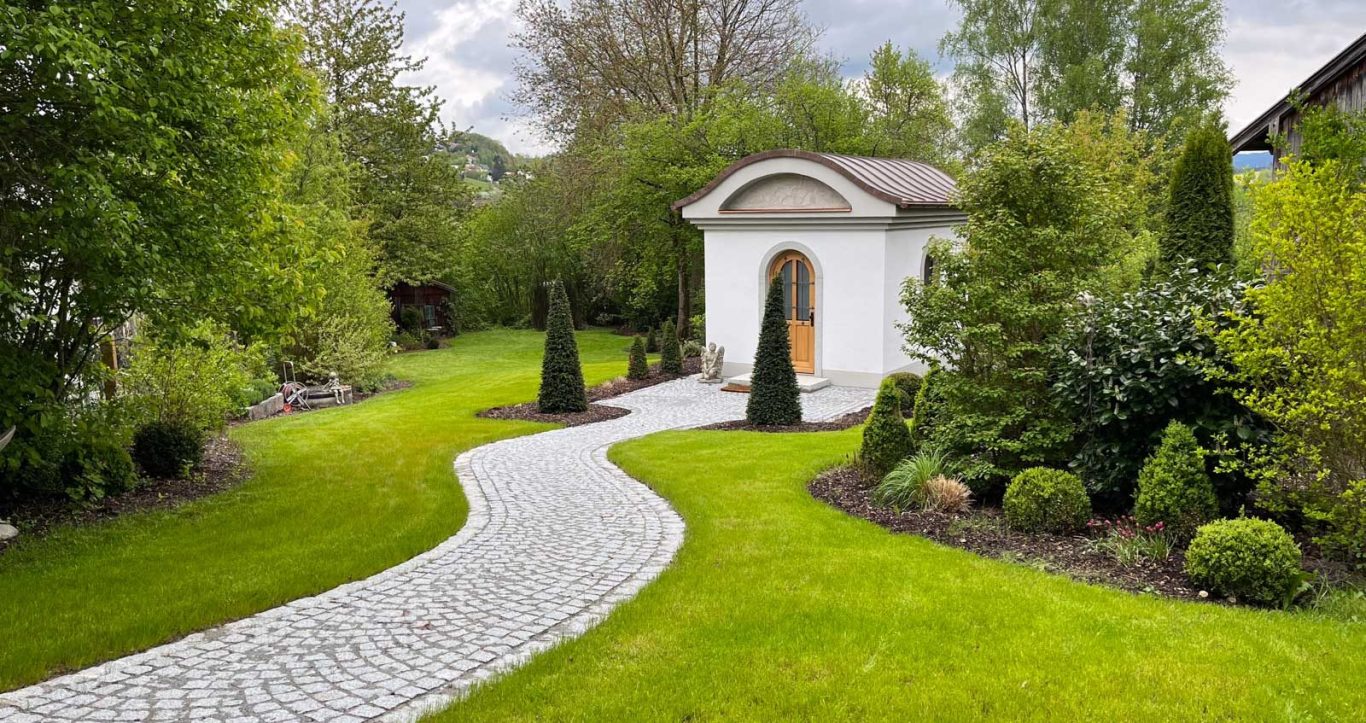 Gepflasterter Weg führt zu einer weißen Kapelle, umgeben von üppigem Grün und Bäumen.