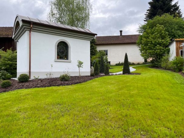 Ein gepflegter Garten mit einem kleinen weißen Gartenhaus und grüner Rasenfläche.