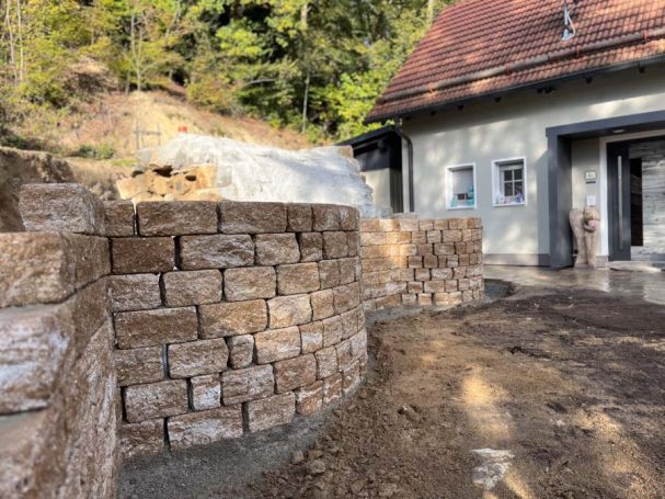 Mauer aus Natursteinen neben einem Gebäude, umgeben von Erde und Bäumen.