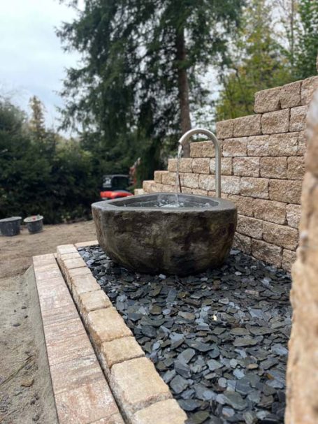 Ein runder, grauer Brunnen aus Stein mit Wasser, umgeben von Kies und einer natürlichen Umgebung.