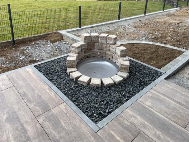 Kreisförmiger Feuerplatz aus Steinen, umgeben von Schotter und Holzplanken.