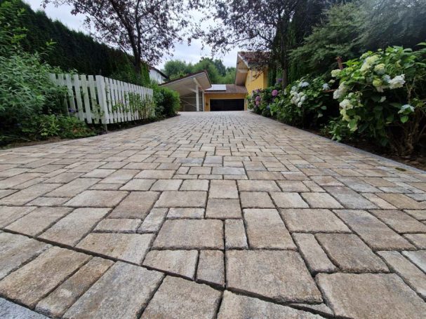 Pflastersteine in einer Einfahrt, umgeben von Bäumen und blühenden Sträuchern.